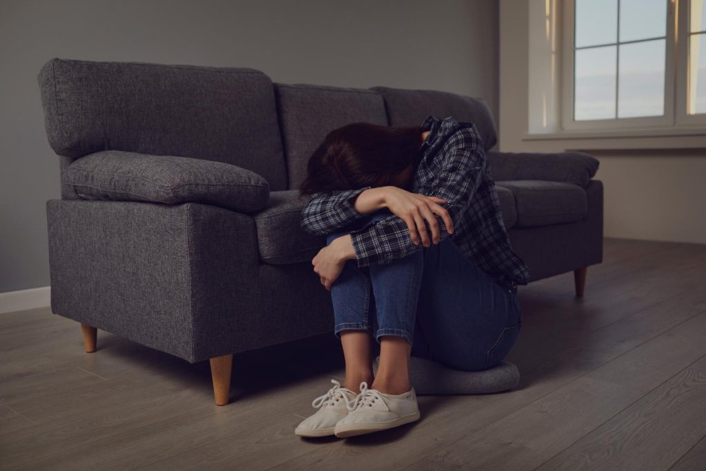 how to stop feeling depressed - a lady sitting on the floor in front of a sofa with her arms around her legs and her head resting on her arm