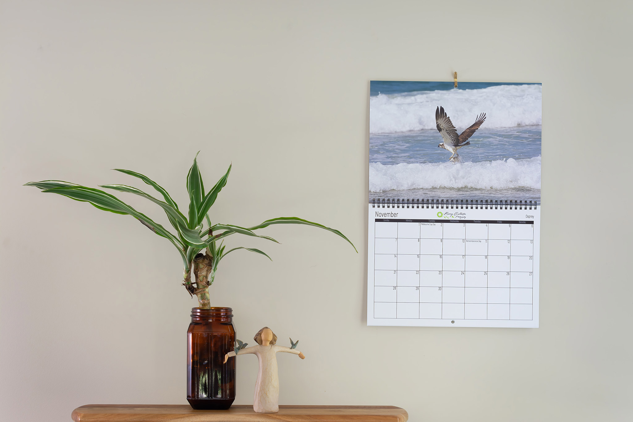 photo of the month September 2020 a calendar hanging on a wall showing a photo of an Osprey catching a fish.  A shelf with a plant in a jar and a small figurine sit to the left of the calendar